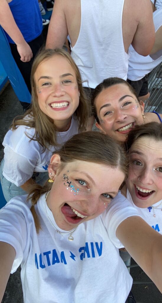 4 girls smiling at the camera, wearing UVic tshirts with UVic colours (blue and yellow) sparkled on their faces.